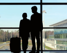 Silhouette of two individuals standing near a window, with one holding a suitcase, suggesting travel or departure